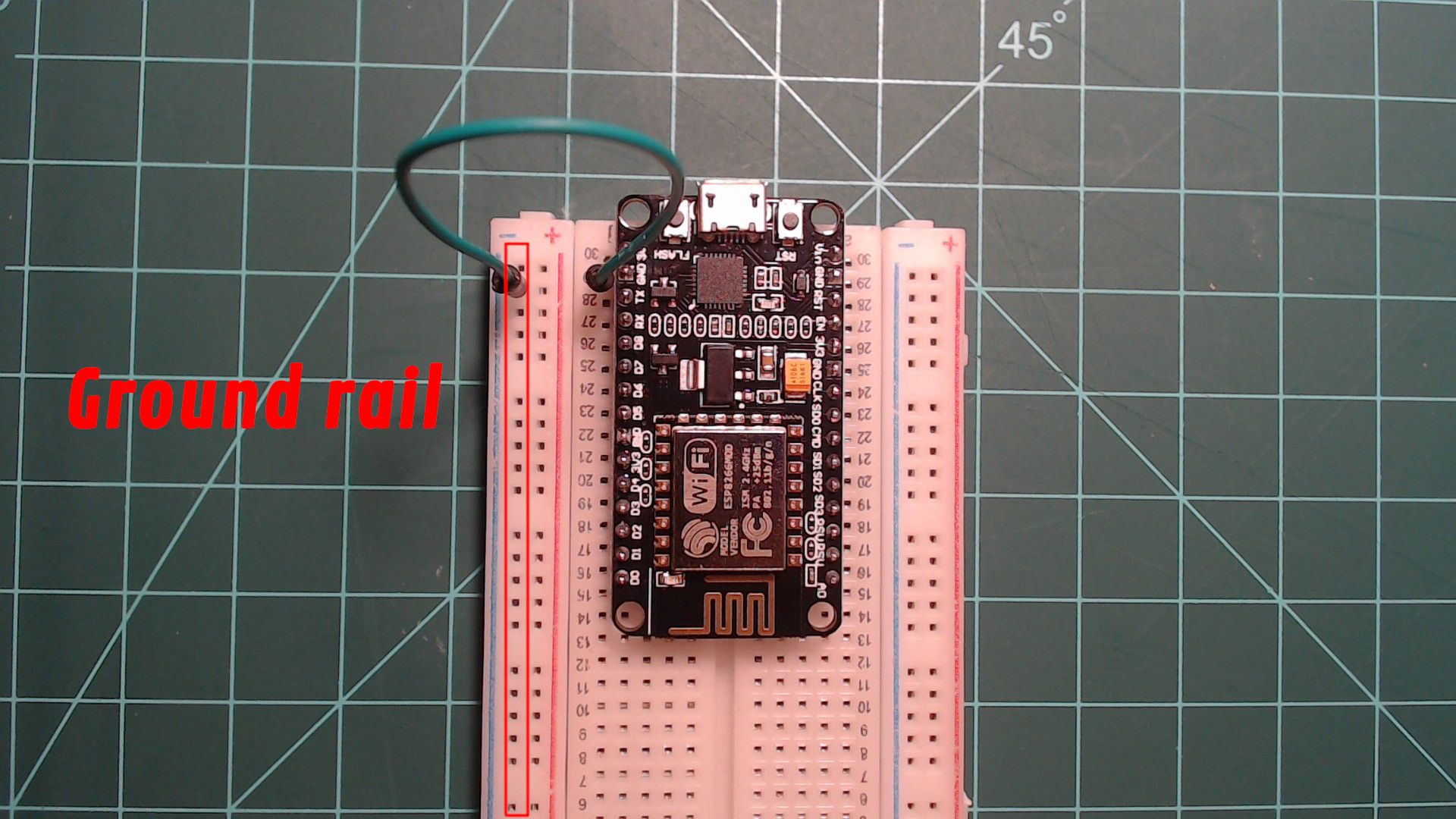 The NodeMCU on the breadboard with a green jumper wire from its GND pin near the USB port to the outermost rail on the left, which is outlined in red and labelled 'Ground rail'.
