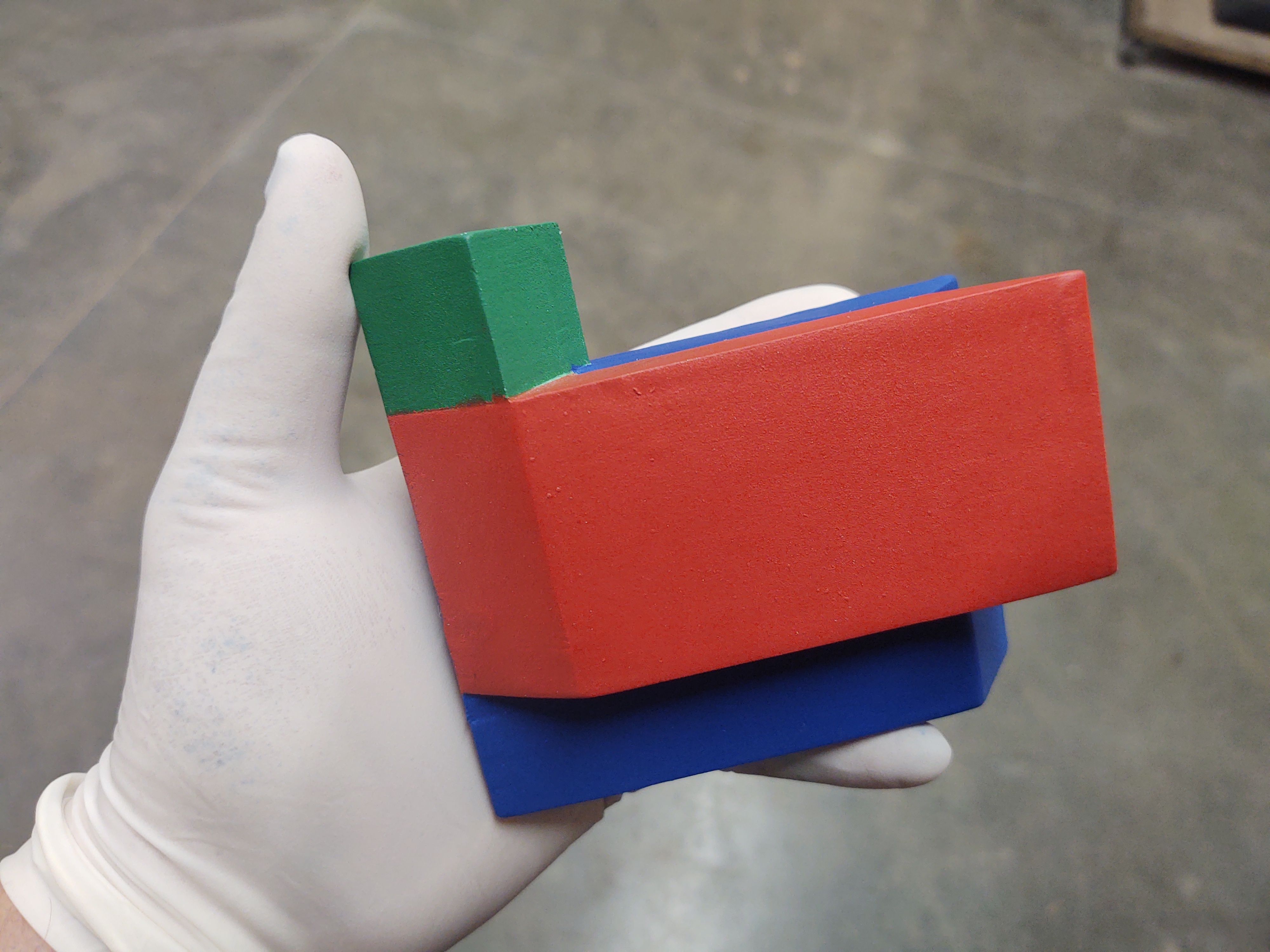 The model held in a gloved hand: the cube on top painted green, the front box red and the back box blue, all with a flat, matte surface.