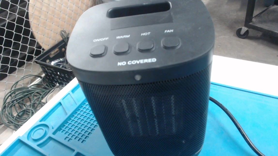 A black space heater on a blue work mat. Its top has a handle slot and four buttons labelled ON/OFF, WARM, HOT and FAN above the printed words NO COVERED, and its front is a black mesh.