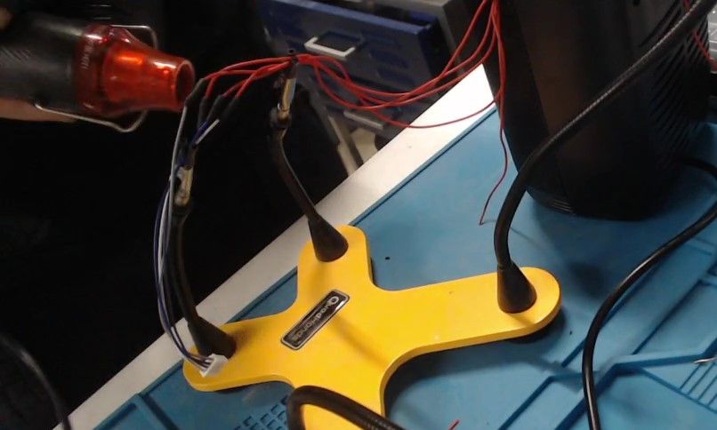 A helping-hands stand holds the red button wires, now ending in a small white connector, while a heat gun is aimed at them from the left.