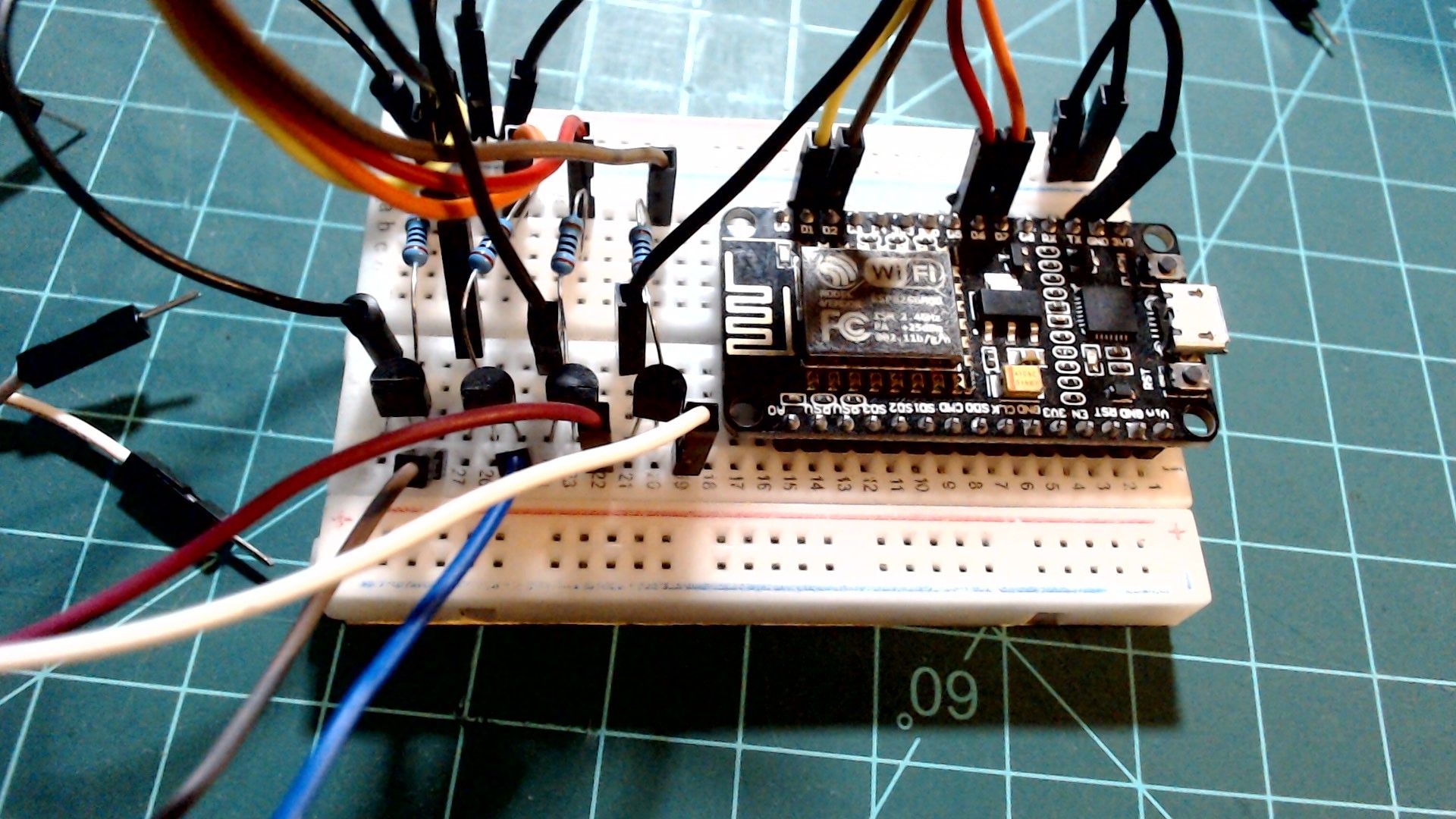 A small breadboard with a NodeMCU on the right and four black transistors in a row on the left, each with a blue resistor above it. Jumper wires run from the NodeMCU's pins to the resistors, and white, blue, grey and dark red wires lead away from the transistors towards the heater.