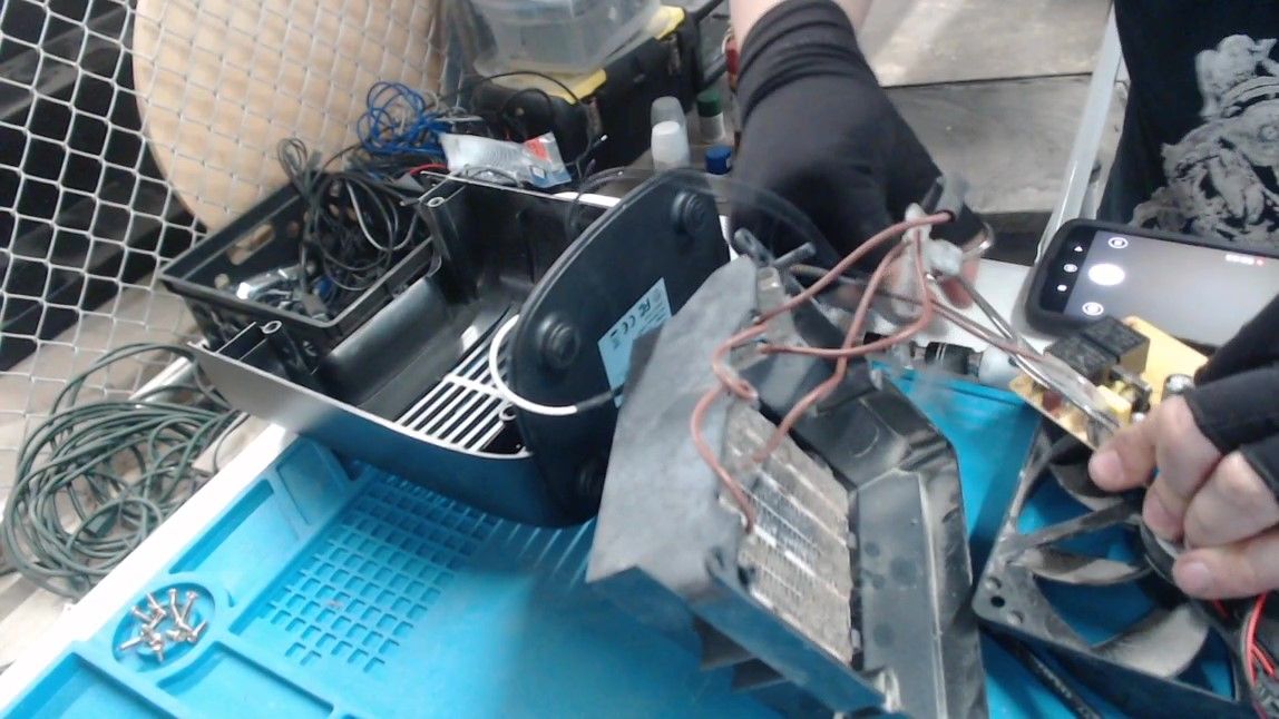 One gloved hand holds the heating element by its brown wires while the other pulls a connector off the circuit board. A phone on the bench has its camera open.