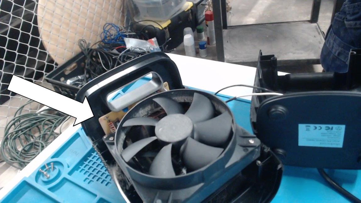 The heater with its back panel lifted off and set aside. A large black fan fills the opening, and a white arrow points to the edge of a circuit board just visible behind it.