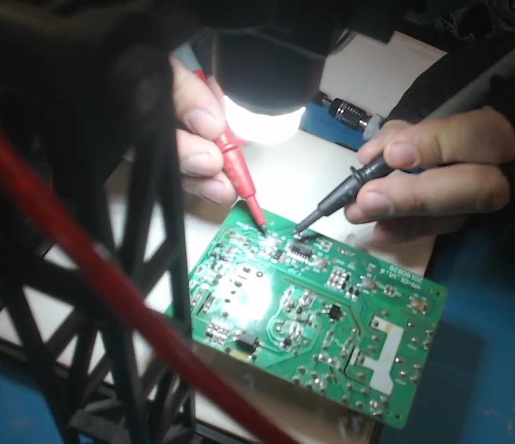 Under a lamp, the red and black probes of a multimeter touch two points on the circuit board's green side.