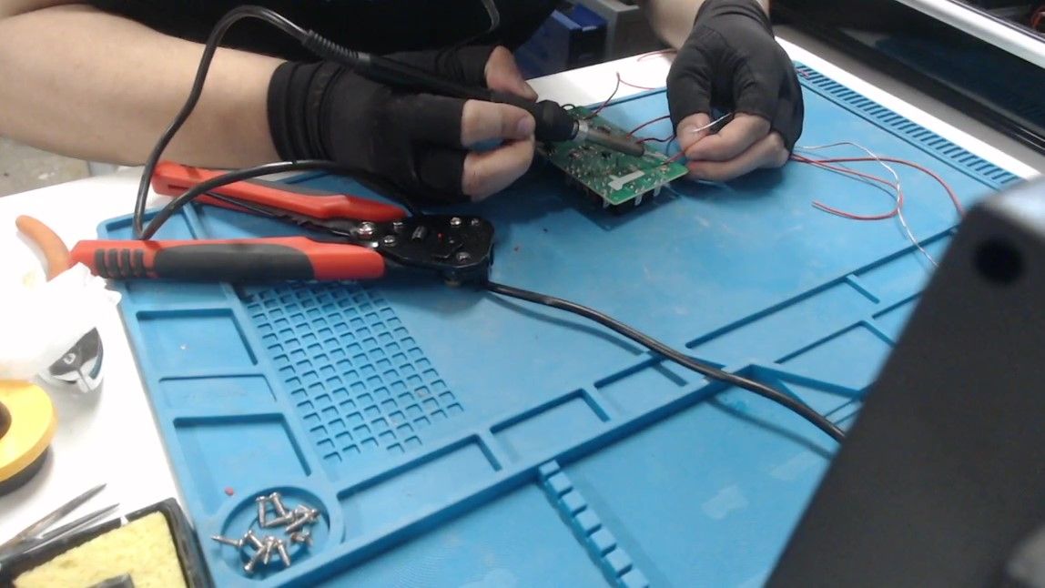 On a blue silicone mat, gloved hands hold a red wire against the circuit board and solder it with a soldering iron, with several other red wires already attached.