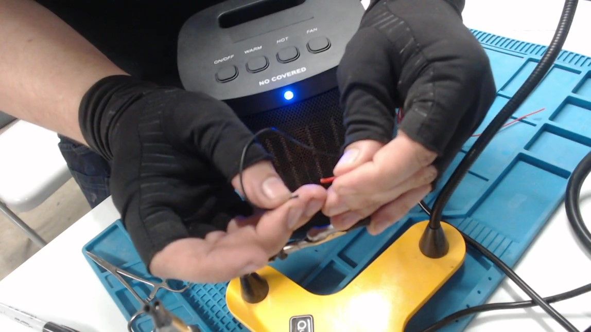 Gloved hands touch the bare end of a red wire to the black wire, and a blue light is lit on the front of the heater below its buttons.