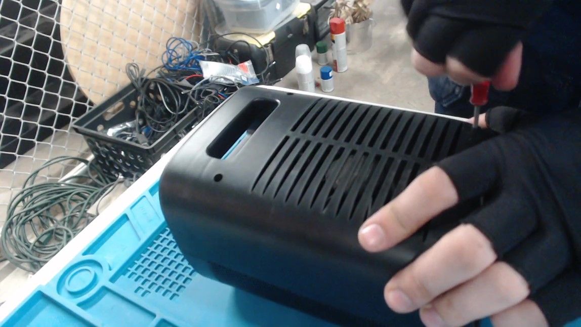 The heater from behind, its vented back panel facing up, while gloved hands undo a screw in the panel with a red-handled screwdriver.
