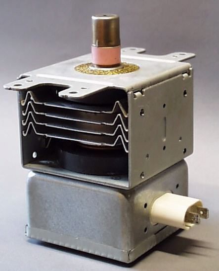 A microwave oven's magnetron: a grey metal block with a stack of thin cooling fins in an open frame, and on top a metal cap standing on a pink ceramic ring.