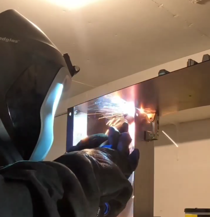 A person in a welding helmet and heavy gloves welds the corner of a steel panel, sparks flying from the joint, with a clamp holding the panel in place.