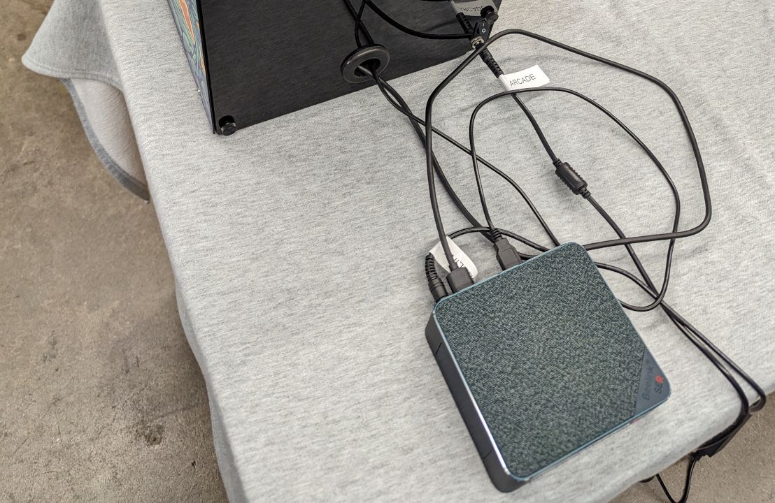 A small square mini PC with a grey fabric top sits on a tablecloth behind a cabinet, its cables running up through a grommet in the cabinet's back panel; a power cable is tagged 'ARCADE'.
