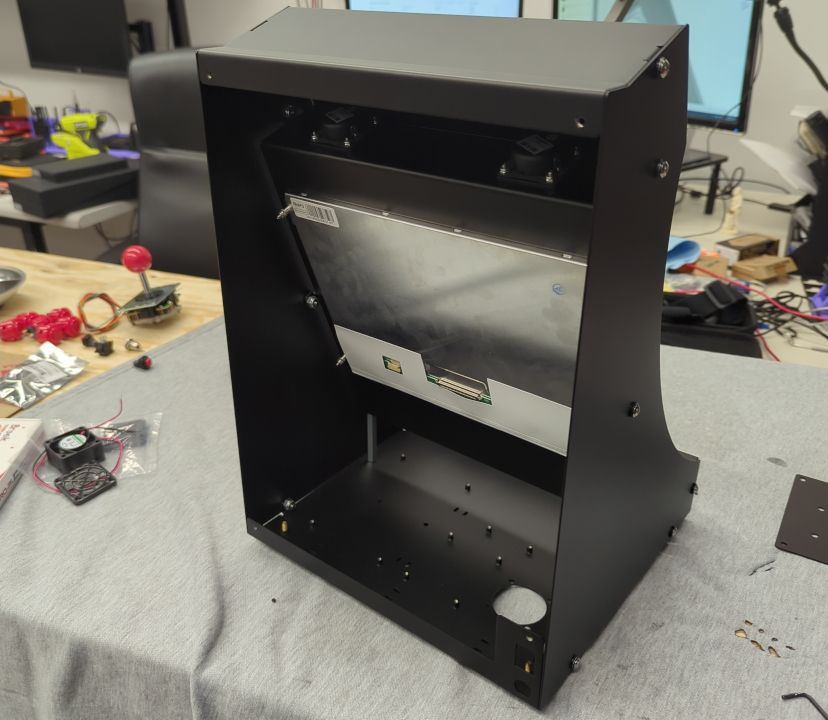 A black powder-coated cabinet, seen from the back with no back panel, standing empty on a workbench: the screen's metal back and two speakers are mounted at the top, and the floor is drilled with mounting holes. Loose joystick, buttons and a fan lie on the bench beside it.
