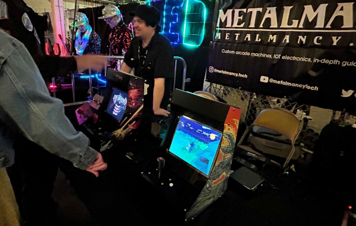 In a dim hall lit by coloured LEDs, a man stands behind two black cabinets with colourful side art, one showing the Thetawave title screen and the other a 3D landscape from the Chickensoft game demo, while a visitor in a denim jacket stands at the front one. The Metalmancy banner hangs at the right.