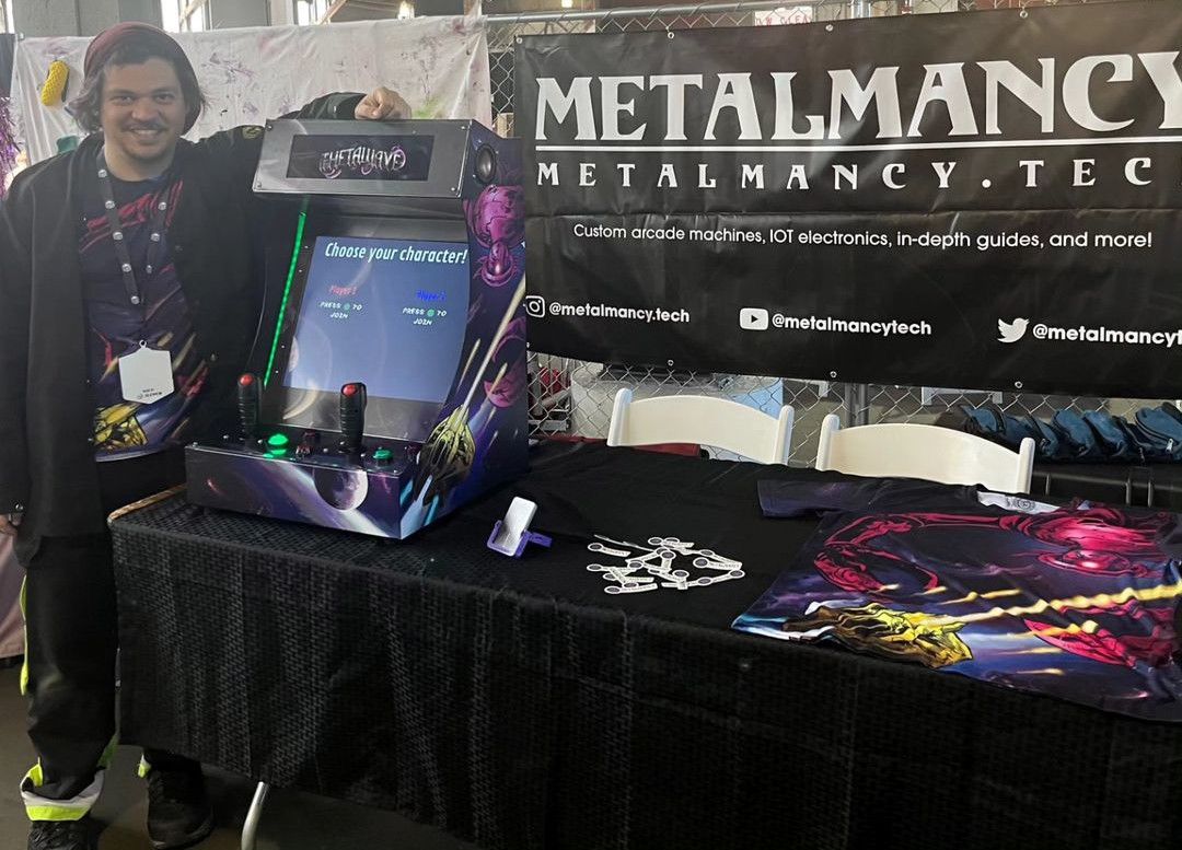 A man in a cap leans on a purple, space-themed cabinet with two joysticks, its main screen reading 'Choose your character!' and a smaller screen above showing the Thetawave logo. Behind the table hangs a black Metalmancy banner; a shirt printed with the same art lies on the table.