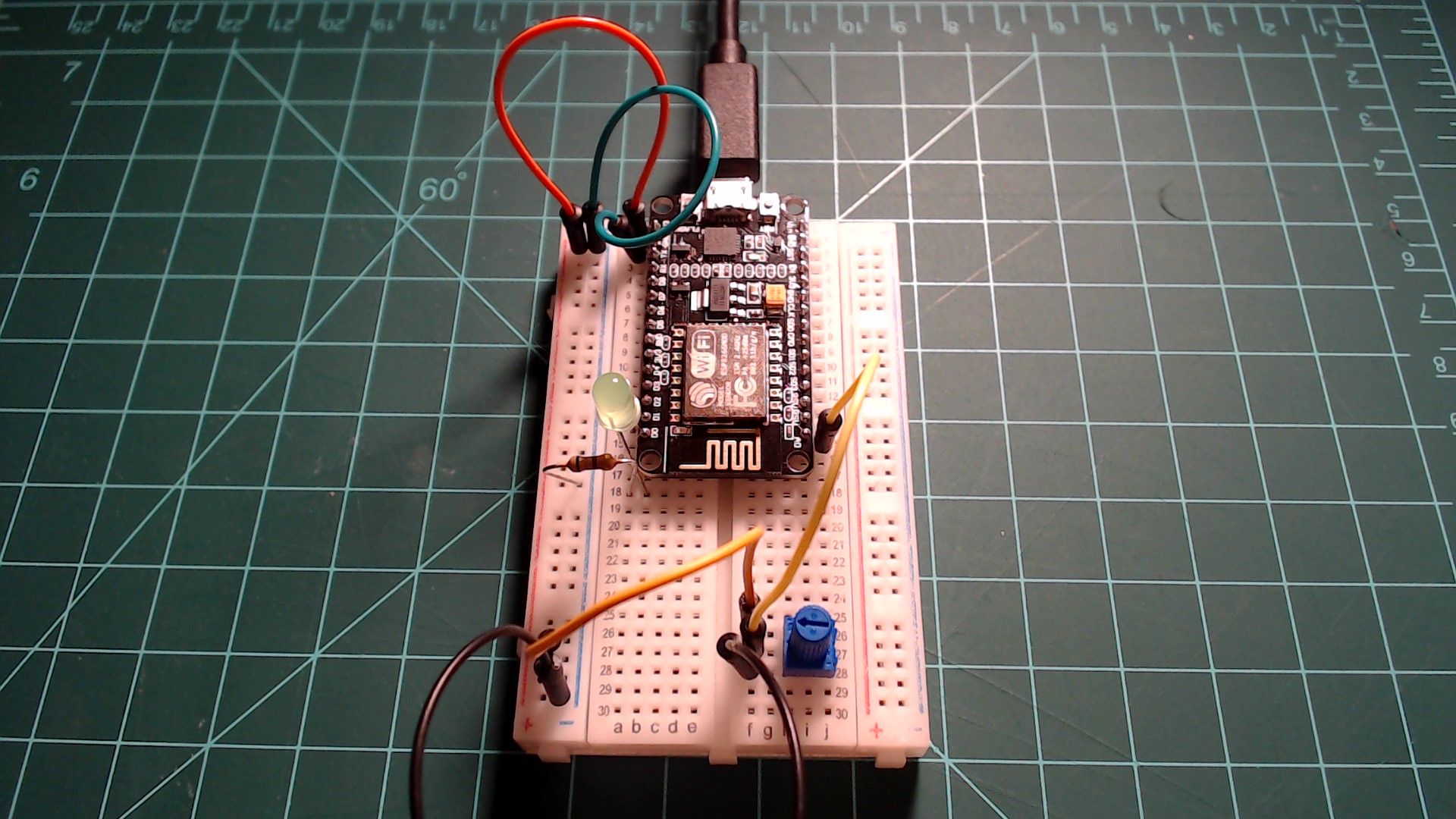 A NodeMCU on a breadboard with a USB cable attached. A red and a green wire run from its 3V3 and GND pins to the rails, an LED and resistor sit beside the board, and yellow and black wires lead to a blue potentiometer near the bottom.