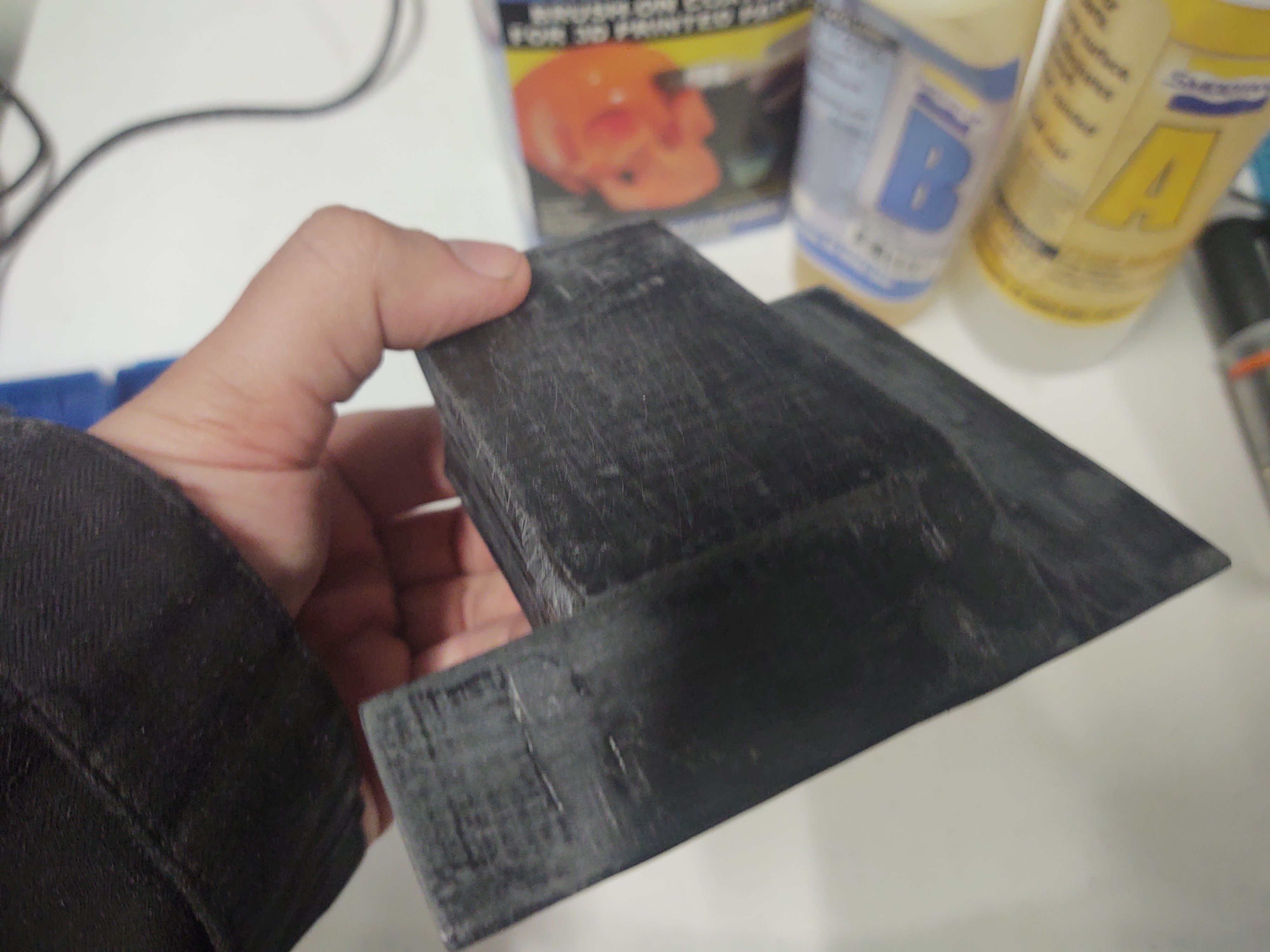 The printed model held in a hand, its surface an even dark grey with a dull finish and fine sanding scratches, no layer lines showing. Behind it stand the coating's box and its two bottles, marked A and B.