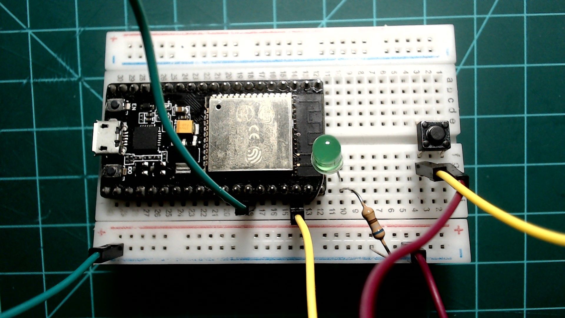 An ESP32 NodeMCU on a breadboard, with a green LED and a resistor beside it in series between GPIO23 and the ground rail, and a push button at the far end wired to GPIO22 with a yellow wire.