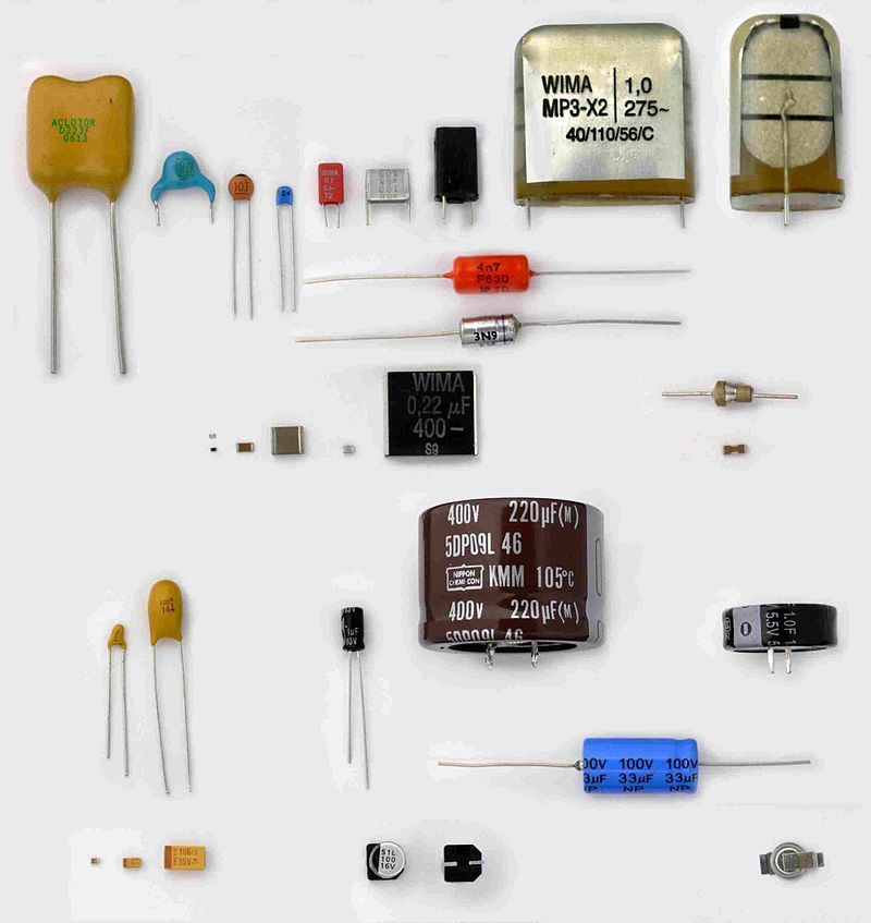 About twenty capacitors laid out on a white background, from tiny surface-mount chips to a large brown 400 V, 220 µF electrolytic can, with yellow, blue and orange ceramic discs, foil film boxes and cylinders with wire leads.