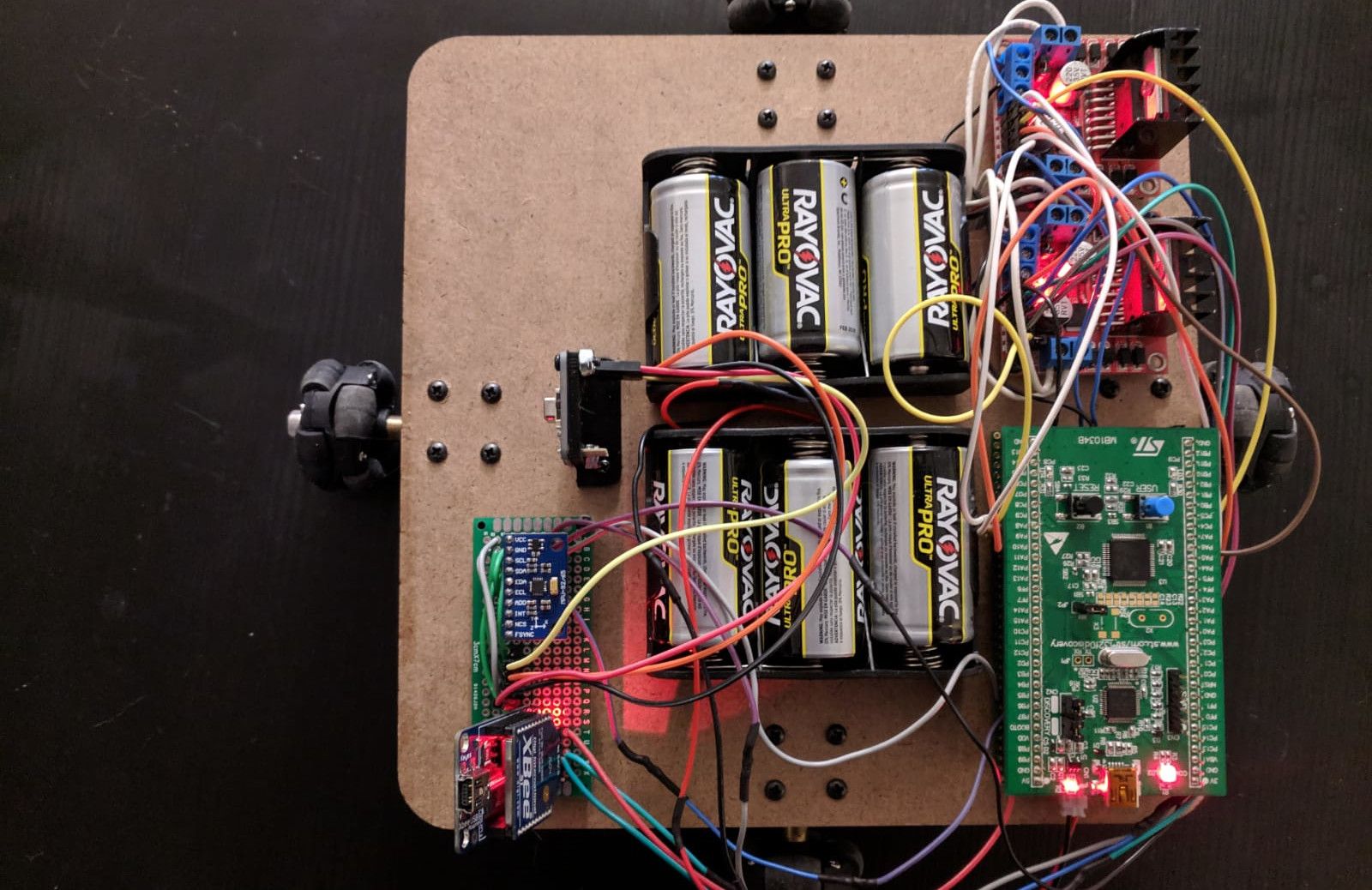 The rover from above: a rounded MDF plate with a roller wheel showing at each side. Two holders of three C batteries fill the middle, the stm32f0discovery board is at the bottom right, the two l298n motor drivers at the top right, and a small protoboard with the imu and the Xbee at the bottom left, all joined by many coloured jumper wires.