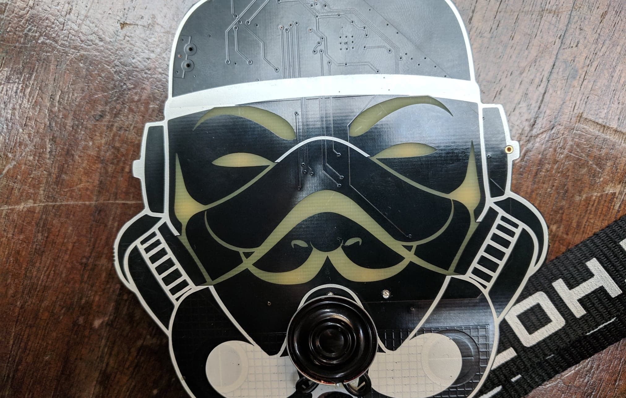 A black circuit board cut into the outline of a helmet, with a white rim, a moustache and pointed beard drawn in pale green across its face, visible copper traces, and a round speaker near the bottom. It rests on a lanyard on a wooden table.