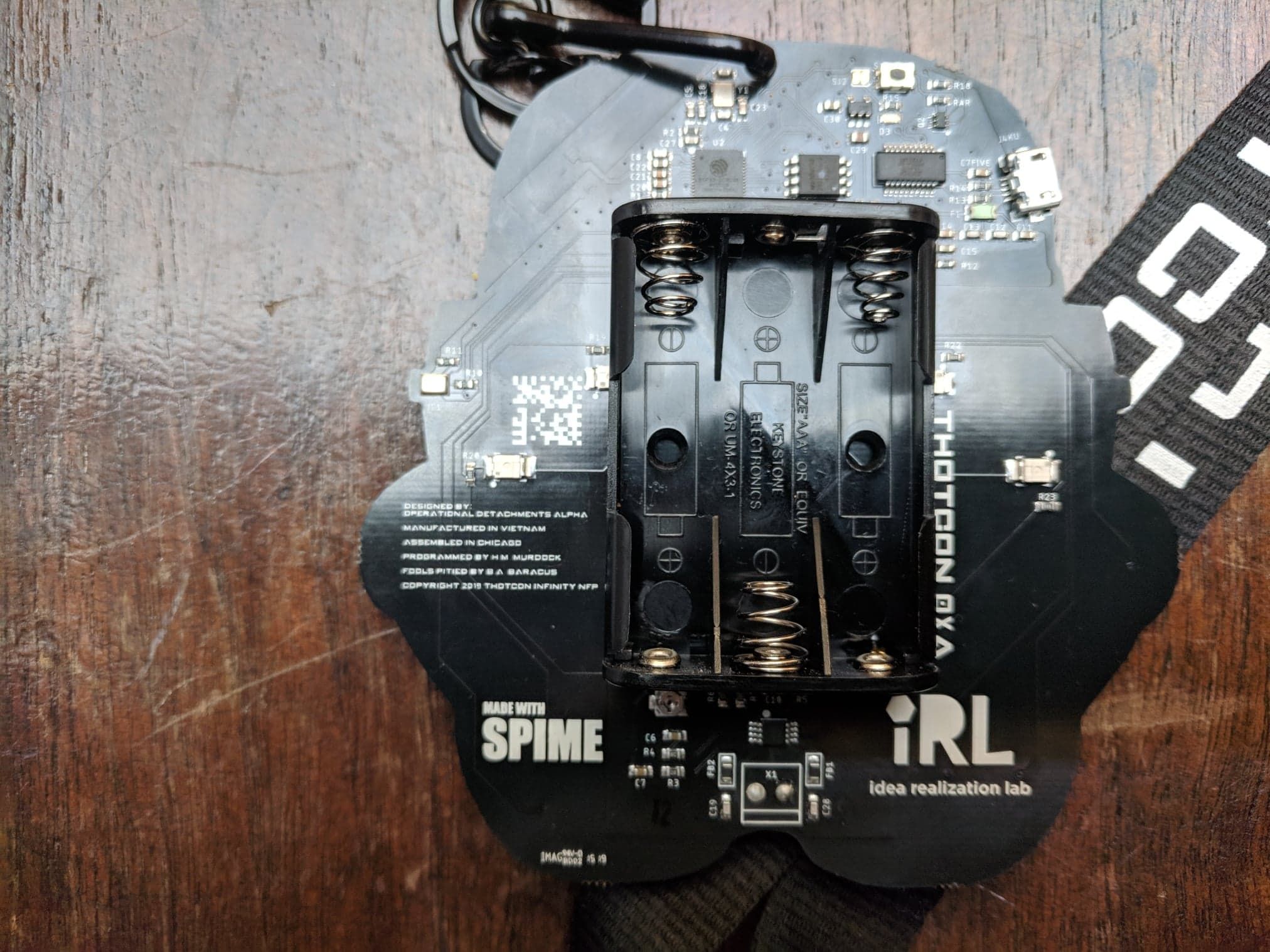 The badge's reverse holds a three-cell AAA battery holder in the middle, with a chip, micro USB port and small components above it. The board is printed 'THOTCON 0XA', 'MADE WITH SPIME', 'iRL idea realization lab' and joke credits such as 'Fools pitied by B.A. Baracus'.