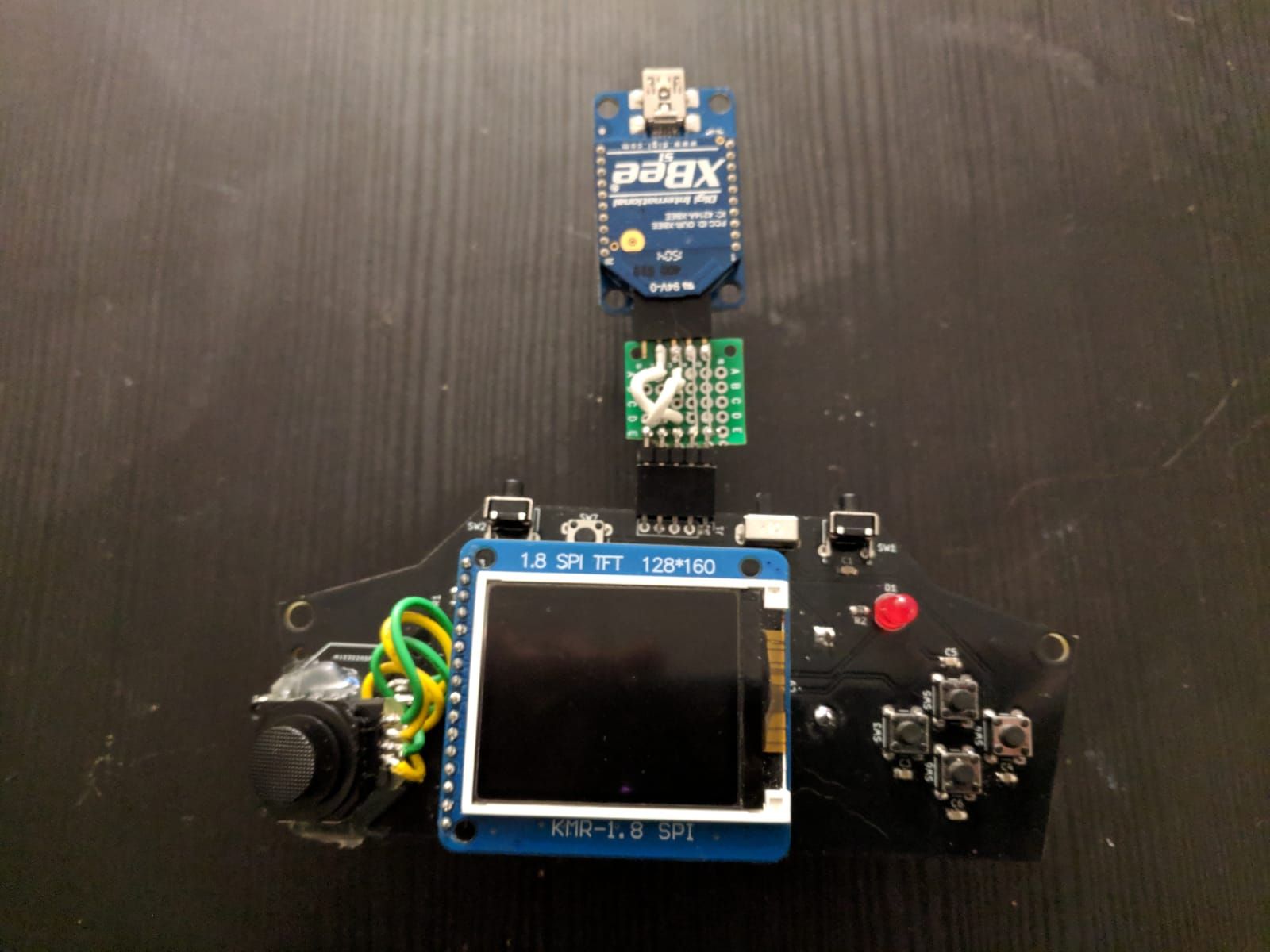 The assembled black board. A 1.8-inch screen module labelled 1.8 SPI TFT 128*160 covers its middle, the PSP analog stick is fixed at the left with yellow and green wires soldered to it, and four buttons sit in a diamond at the right under a red LED. The Xbee, on its blue adapter, plugs into the top header through a small green protoboard on which two white wires cross over.